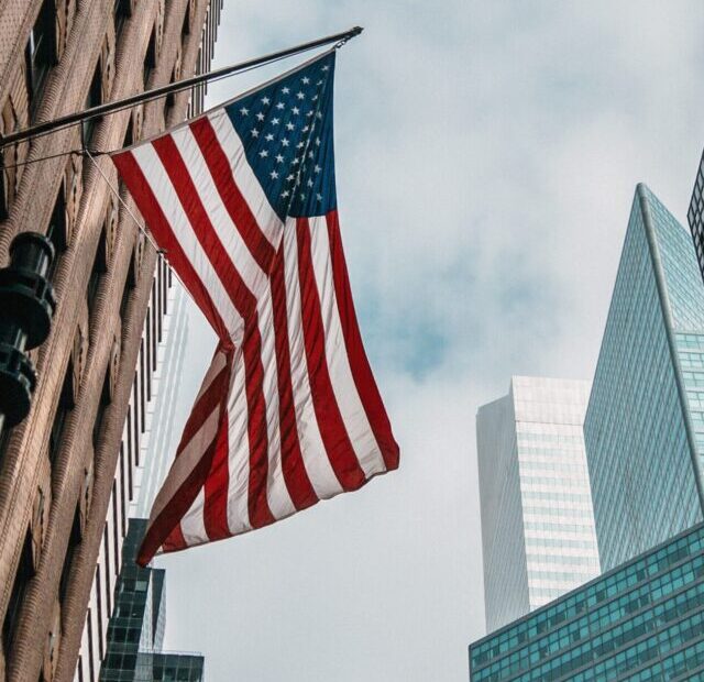 the USA or United States of America flag on a flagpole near skyscrapers under a cloudy sky